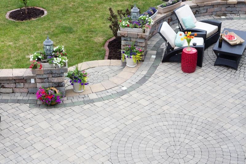 A circular patio features expertly installed paver stones, a black wicker furniture set with white cushions, a red side table, and vibrant flower arrangements along a stone wall.