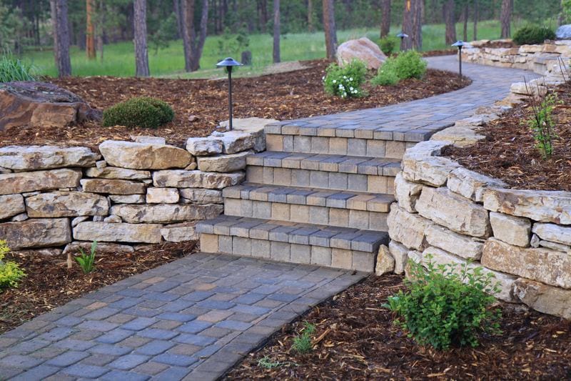 Stone pathway with brick steps flanked by curved stacked stone retaining walls and small garden lights, surrounded by mulched landscaping and greenery. This elegant design showcases the expertise of a skilled hardscape contractor.