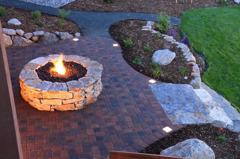 A paver stone fire pit with a lit flame is nestled on a brick patio, surrounded by a landscaped garden adorned with rocks and greenery.