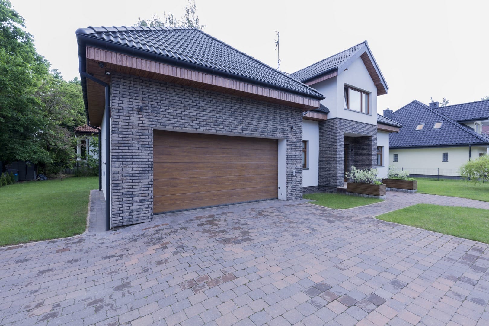 Modern two-story house with grey brick exterior and a large wooden garage door, surrounded by a paved driveway and lawn, expertly crafted by a skilled hardscape contractor.