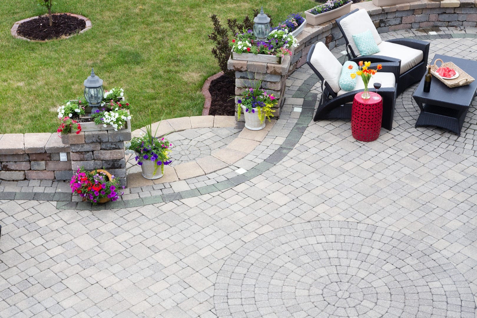 The patio, crafted by a skilled paver stone contractor, boasts circular brick patterns and offers a cozy sitting area with cushions. A red side table complements the scene with vibrant flowers in pots and a platter of watermelon slices on a sleek black coffee table.