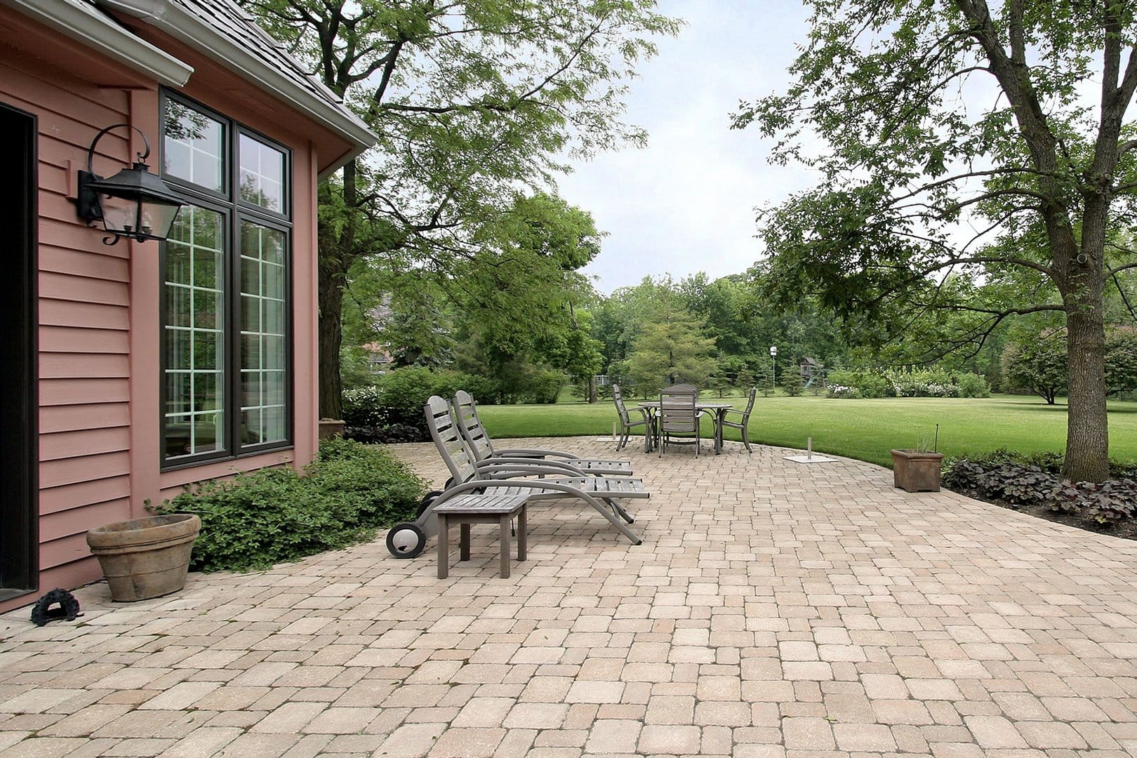 A brick patio featuring lounge chairs and a round table set lies adjacent to a house with large windows, surrounded by trees and lush grass. The stunning design was achieved through interlocking paving installation, highlighting the beauty of expertly placed paver stones.