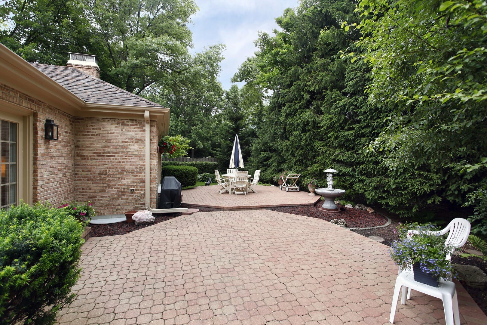 A charming paver stone patio with outdoor furniture, an umbrella, a grill, and a fountain. Enveloped by lush greenery and trees. A white chair with a potted plant graces the foreground, creating a serene setting.